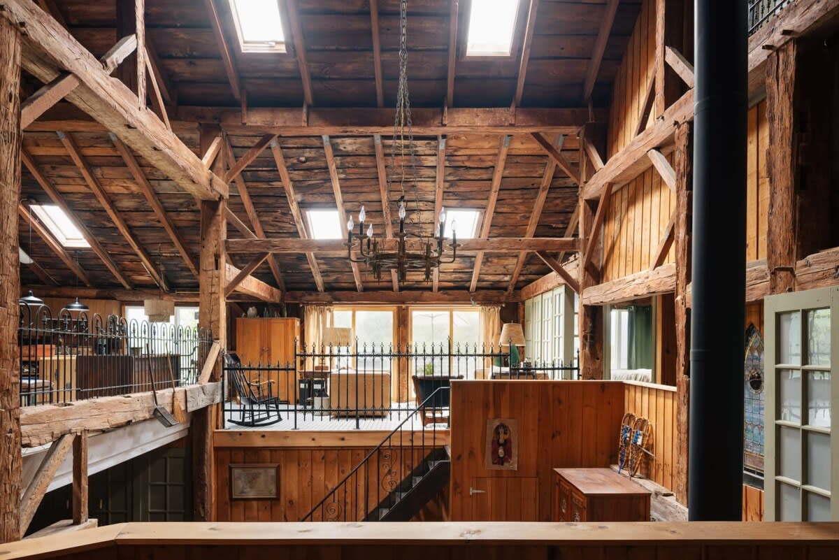 Grand interior view showcasing the barn's vaulted wooden ceiling with massive exposed beams and a large chandelier. A railing overlooks the lower level, and natural light streams in from skylights.