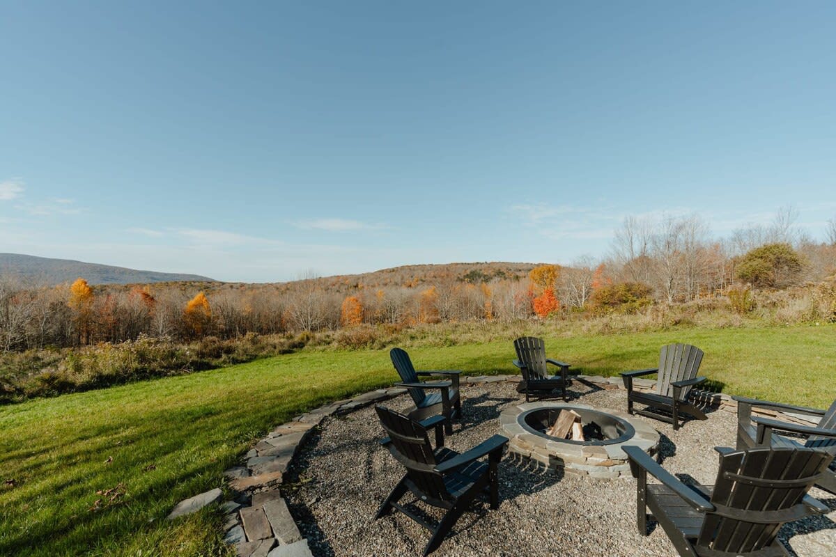 A circular stone fire pit area with dark Adirondack chairs is nestled on a green lawn, offering panoramic views of distant hills adorned with vibrant autumn foliage under a clear blue sky.