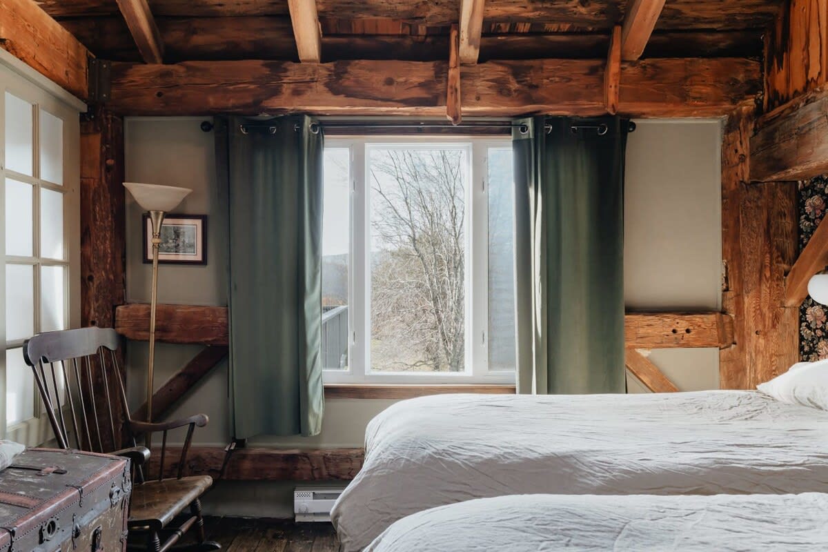Bright bedroom corner showcasing a large window with green curtains, offering views of the outdoors. A wooden rocking chair and a vintage trunk sit on the light hardwood floor, complementing the rustic exposed beams.