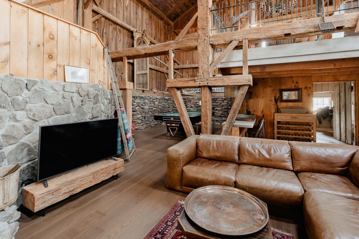 Expansive living area with a large leather sectional sofa and a flat-screen TV on a rustic wooden stand, transitioning into a game area with a pool table in the background. The space features stone walls and exposed wooden beams.