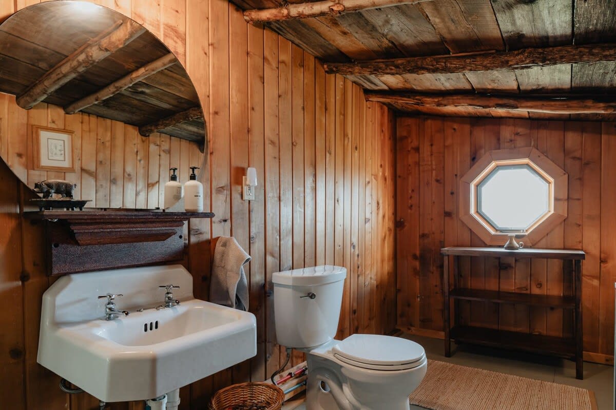 Rustic bathroom featuring warm wood-paneled walls and ceiling, a white pedestal sink with a round mirror, and a white toilet. A unique octagonal window adds character to the space.