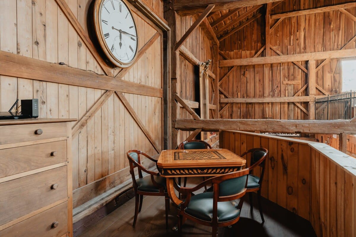 Upper-level game area with a small wooden card table and four chairs, set against warm wooden walls and exposed beams. A large round clock adorns the wall, adding a classic touch to the recreational space.