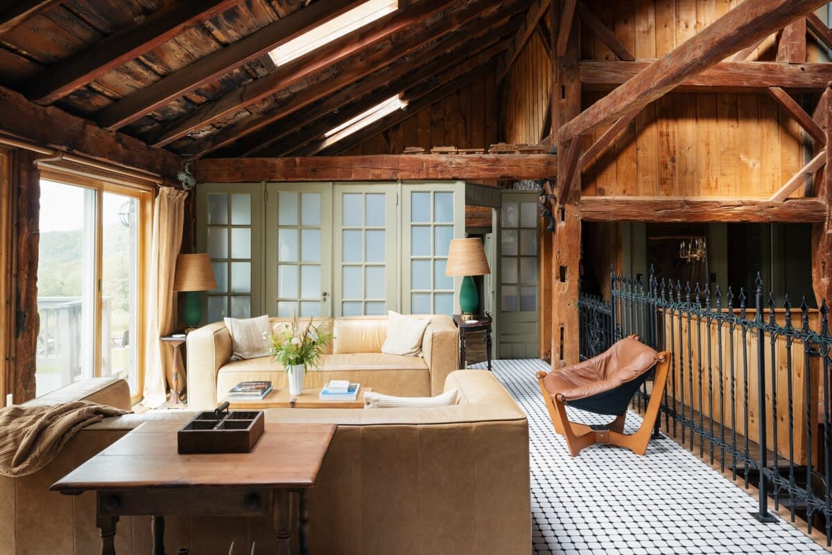Cozy living area with two light brown leather sofas, a wooden coffee table, and large windows offering natural light. The room features a vaulted wooden ceiling with exposed beams and a black and white tiled floor.