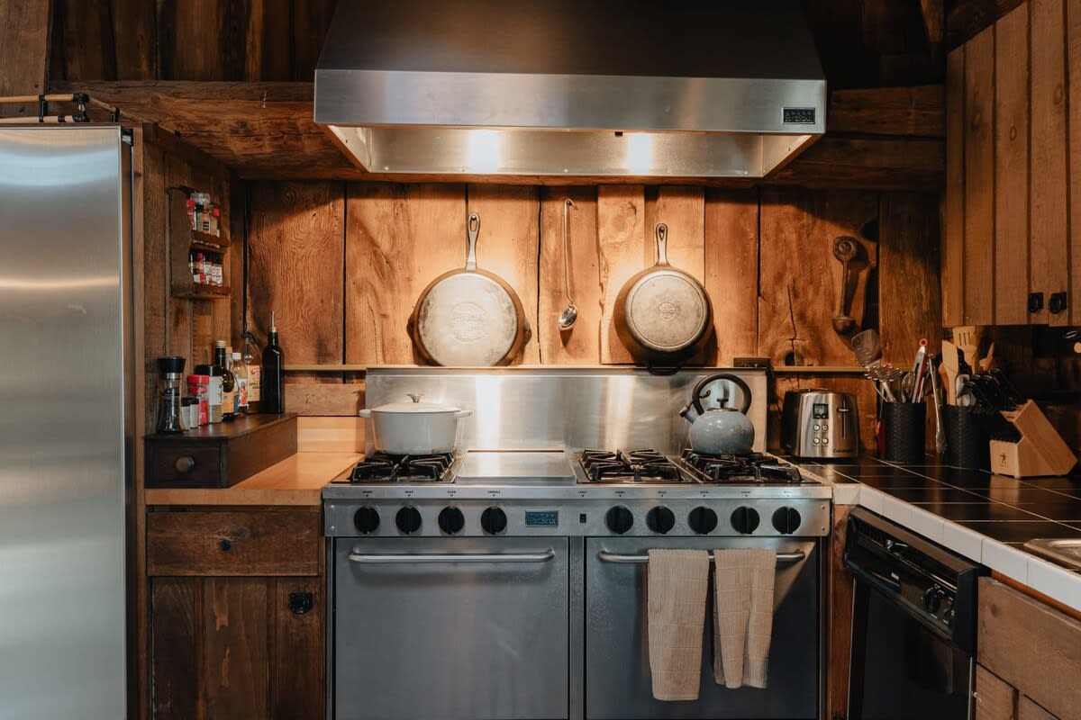 Close-up of the kitchen's stainless steel professional range and hood, set against a rustic wooden wall with hanging cast iron cookware. The counter space includes a kettle and toaster.