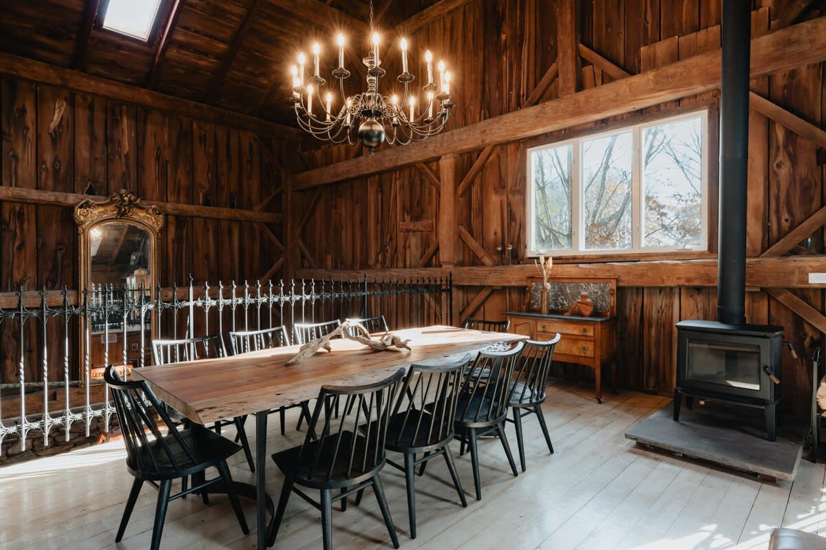 A spacious dining area features a long, rustic wooden table with black spindle-back chairs, illuminated by an ornate chandelier. The room is characterized by dark wood paneling, exposed beams, and a large window offering natural light.