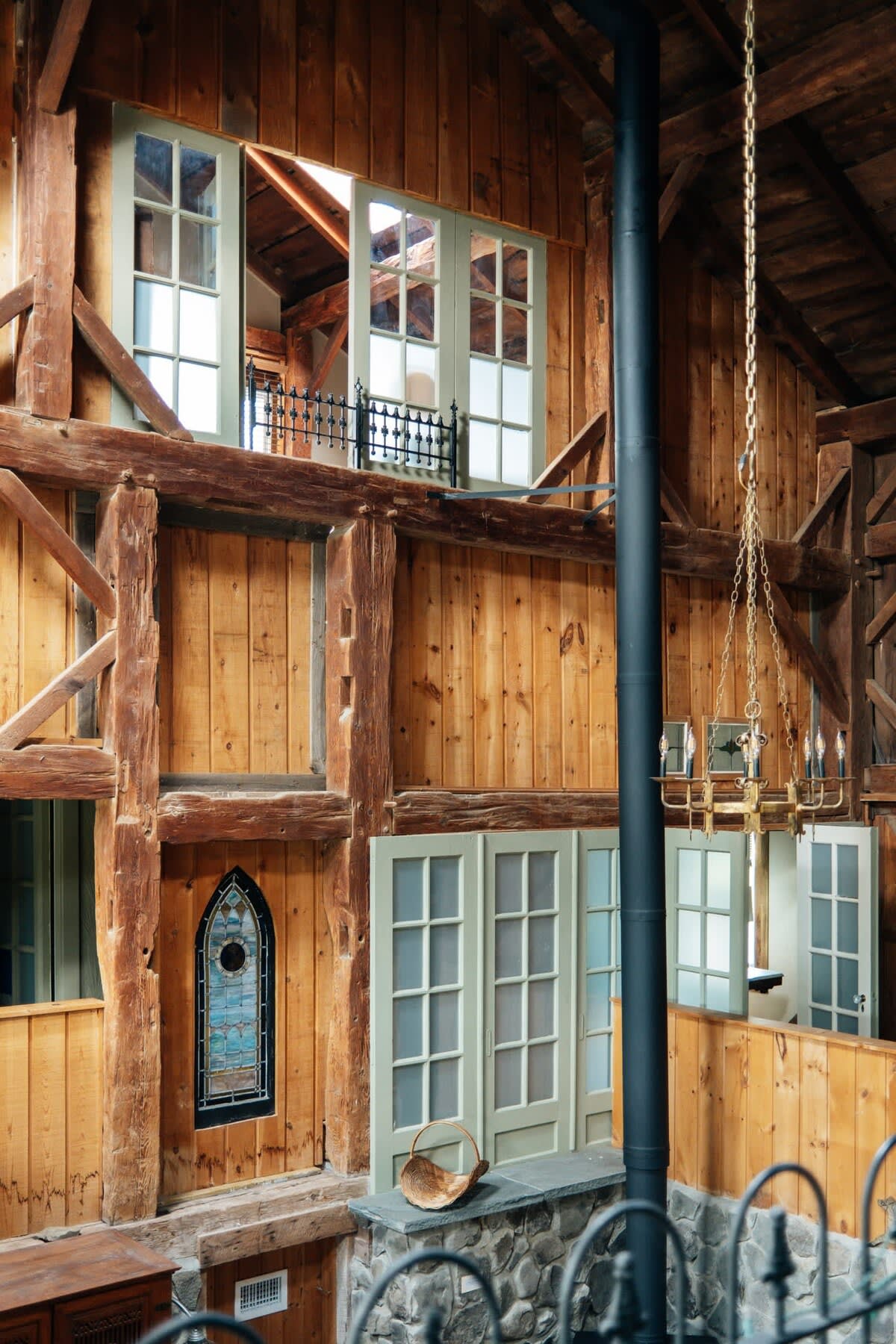 Dramatic interior view highlighting the barn's tall wooden walls and exposed timber framing, with multiple windows and a unique arched stained-glass window. A black stovepipe runs vertically, emphasizing the height.