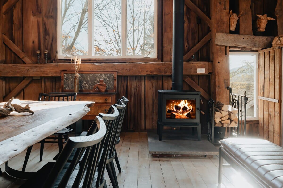 Rustic dining area featuring a long, live-edge wooden table with black chairs, set against a backdrop of warm wooden walls and a large window. A black wood-burning stove with a visible fire adds a cozy focal point.