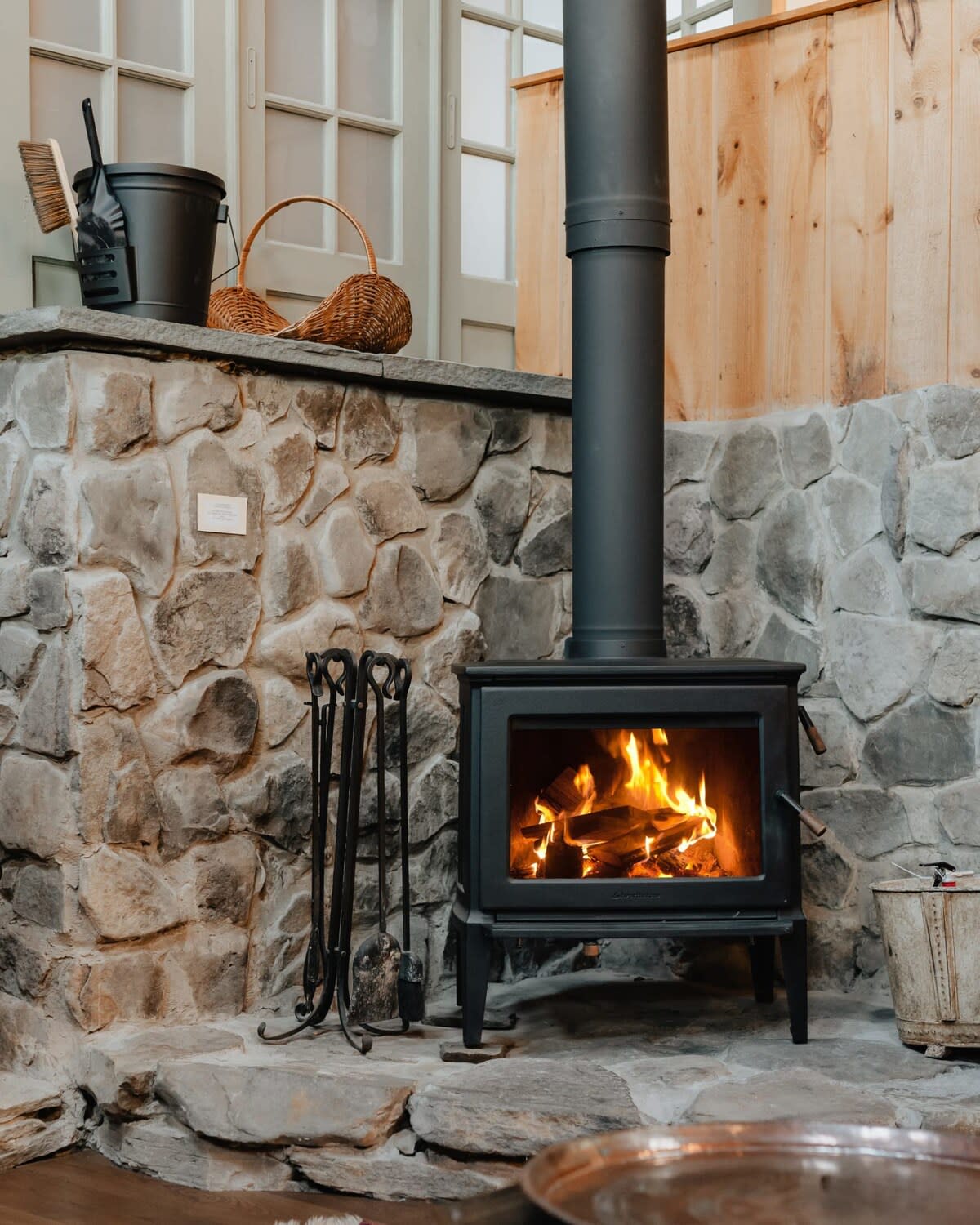 Close-up of a black wood-burning stove with a roaring fire, set on a stone hearth against a backdrop of natural stone walls. Fireplace tools and a wood basket are neatly arranged beside it.