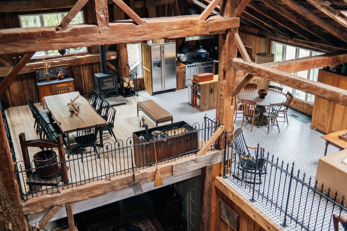 An expansive open-concept living area, viewed from above, showcases a kitchen with stainless steel appliances, a large dining table, and a round breakfast nook, all within a rustic barn structure with exposed wooden beams and white tiled floors.
