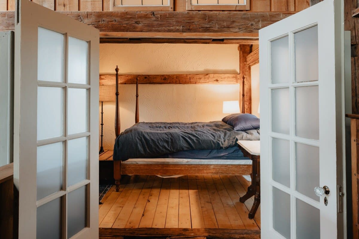 View into a charming bedroom through white frosted French doors, revealing a dark wooden four-poster bed with blue bedding. The room features light wooden floors and exposed timber beams, creating a rustic yet inviting atmosphere.