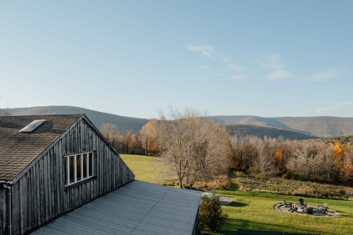 Elevated view of a rustic barn with weathered wooden siding and a shingled roof, featuring a skylight and window, overlooking a green yard with a stone fire pit and chairs, all set against rolling mountains and autumn trees.