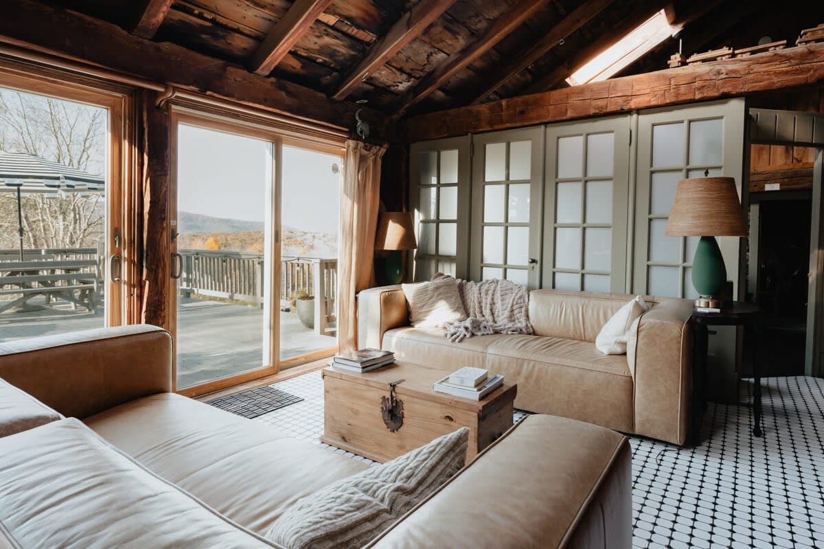 Bright living space with two light brown leather sofas, a wooden coffee table, and sliding glass doors opening to an outdoor deck. The room has a vaulted wooden ceiling and a black and white tiled floor.