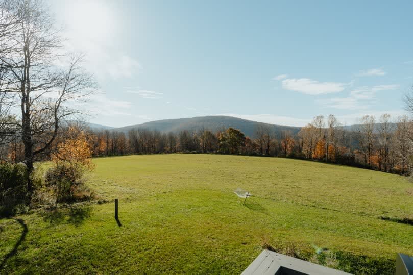 A vast green field stretching towards distant mountains under a bright blue sky, with scattered trees showing hints of autumn colors. A clothes drying rack is visible in the field.