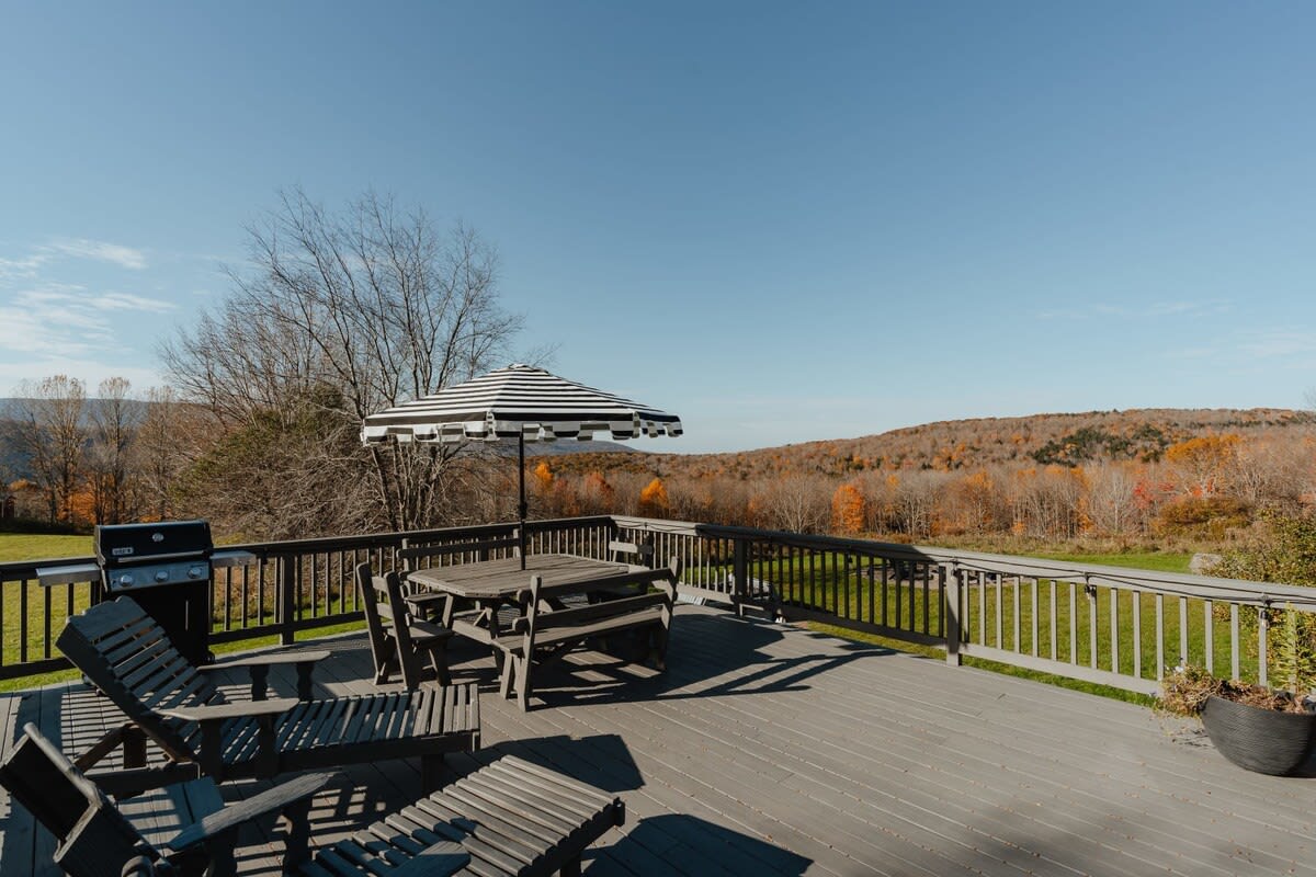 A spacious outdoor deck features a wooden dining set with a striped umbrella, lounge chairs, and a BBQ grill, all overlooking a scenic landscape of rolling hills and autumn trees under a bright blue sky.