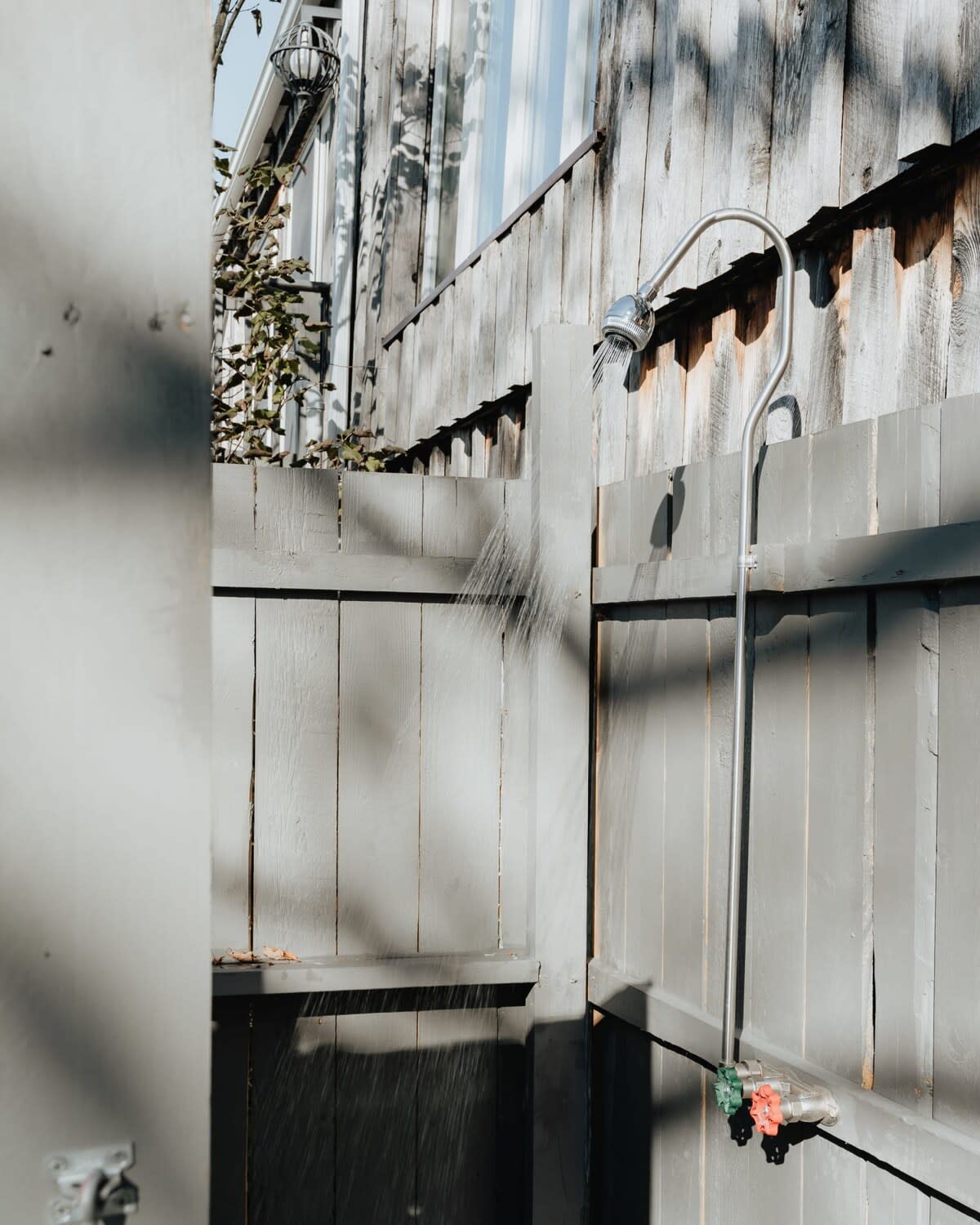 An outdoor shower with gray wooden privacy walls, featuring a chrome shower head and visible water spray. The surrounding fence is also gray.