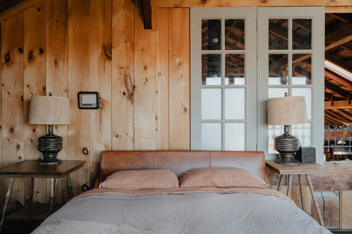 Close-up of a comfortable bed with a light brown leather headboard and neutral-toned bedding, flanked by two rustic wooden nightstands with matching lamps. Wood-paneled walls and a large sliding barn door with glass panes are visible.