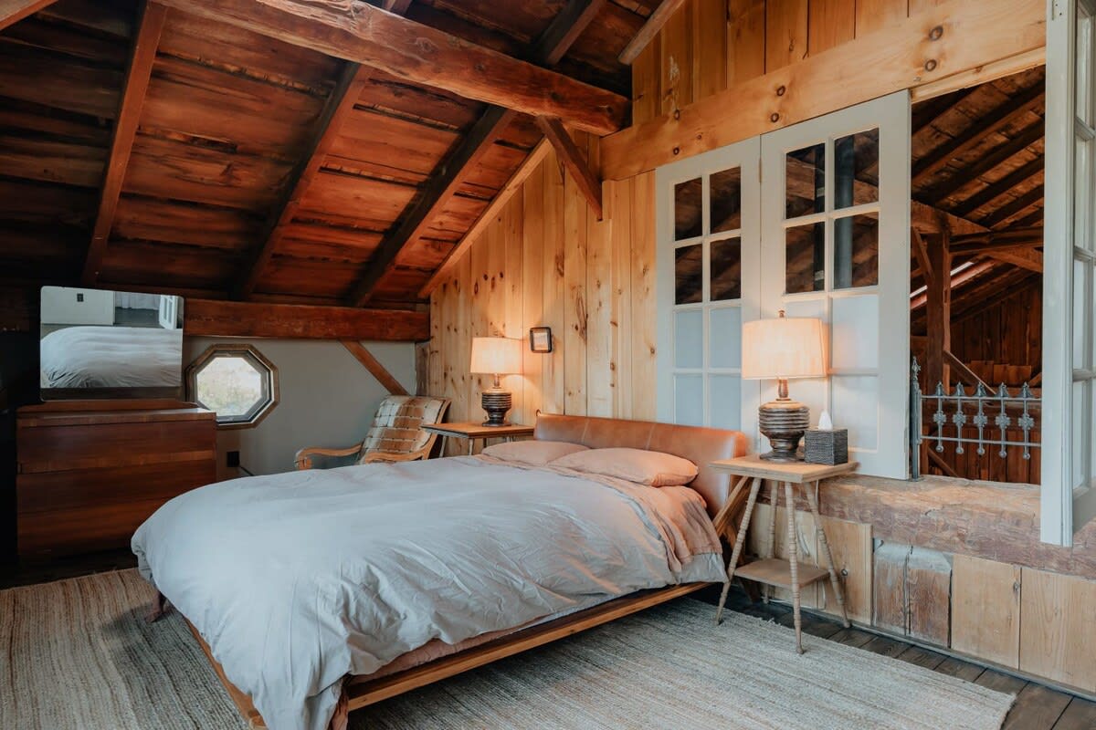 Cozy bedroom with a light-colored bed, two wooden nightstands with lamps, and rustic wood-paneled walls and ceiling with exposed beams. A small octagonal window is visible in the background.