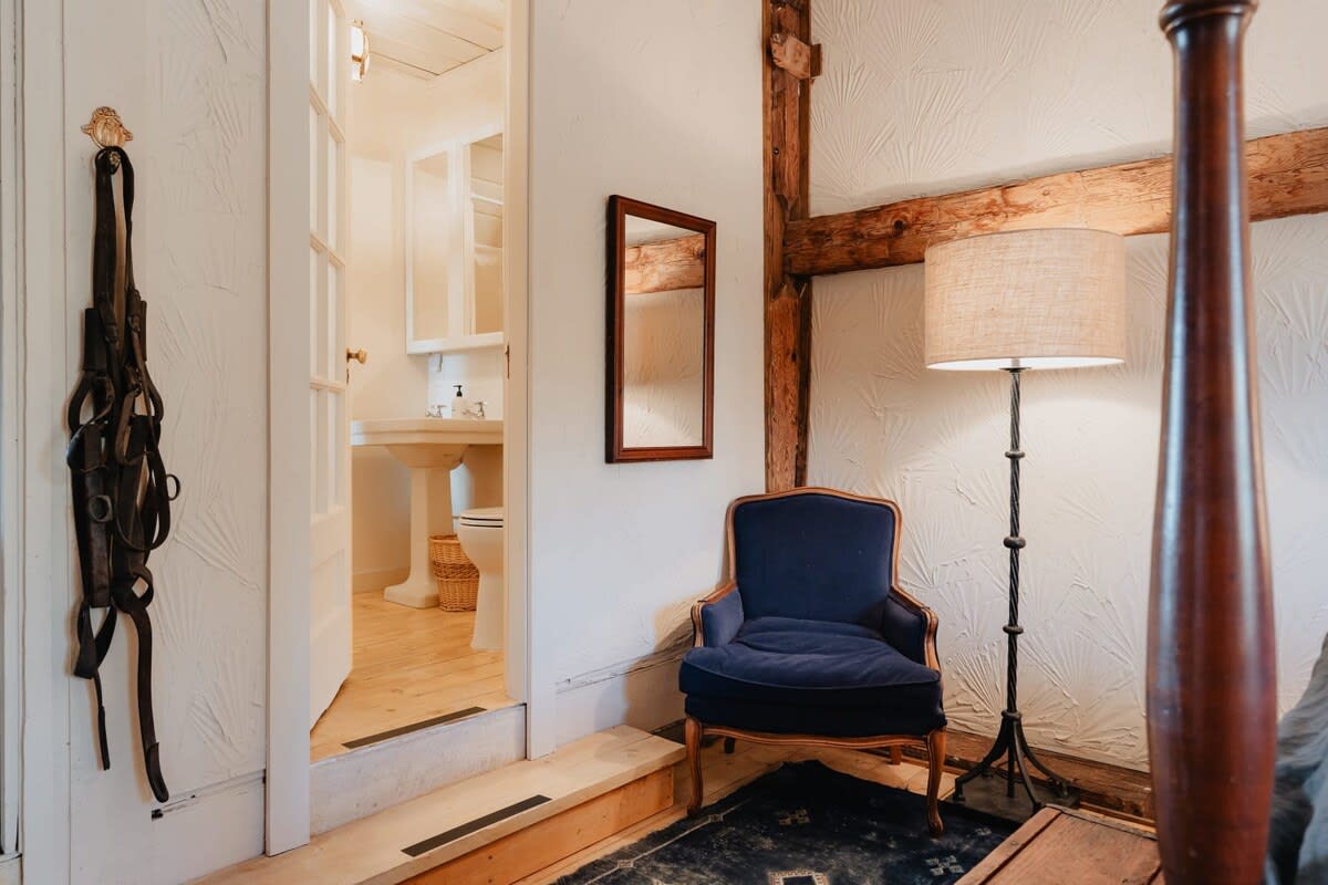 Corner of a rustic bedroom with a dark blue armchair and a tall floor lamp, leading into a bright en-suite bathroom. Exposed wooden beams and textured white walls maintain the barn's character.