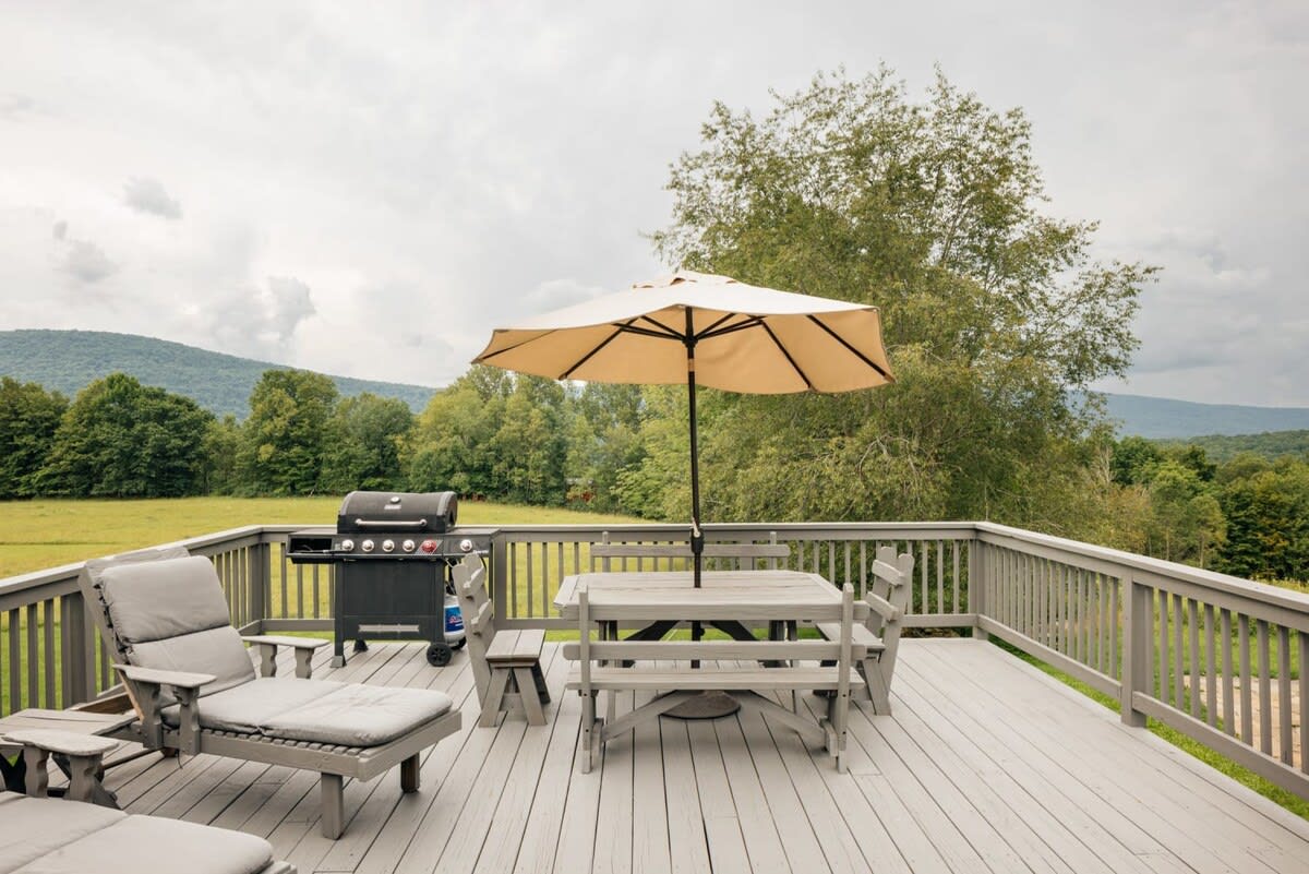 A spacious outdoor deck with a gray wooden railing, featuring a picnic table with an umbrella, lounge chairs, and a BBQ grill, all overlooking a green field and distant mountains under a cloudy sky.