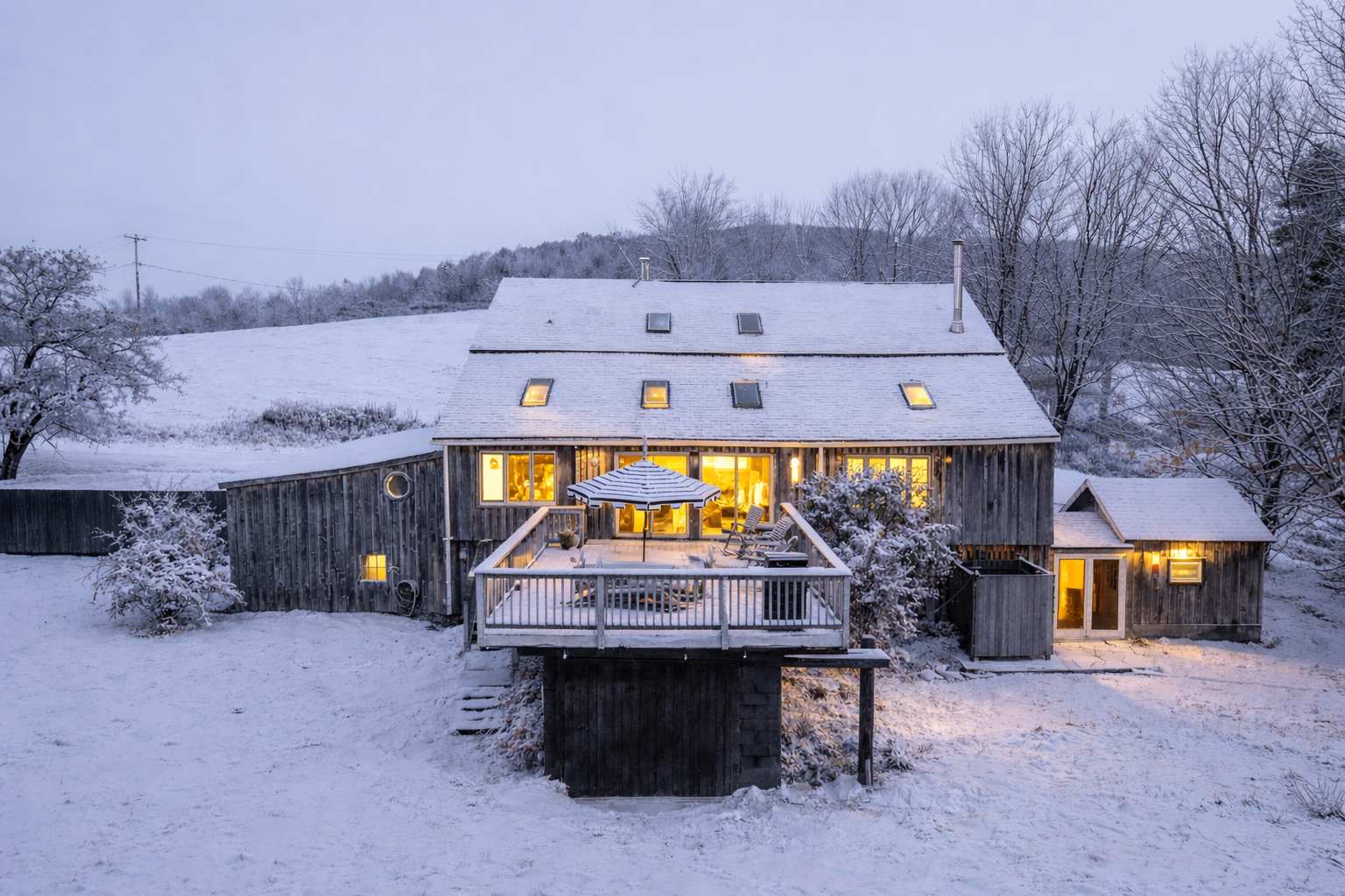 An aerial view of the barn-style house covered in snow during winter, with warm lights glowing from inside and a large deck visible. The surrounding landscape is also snow-covered.