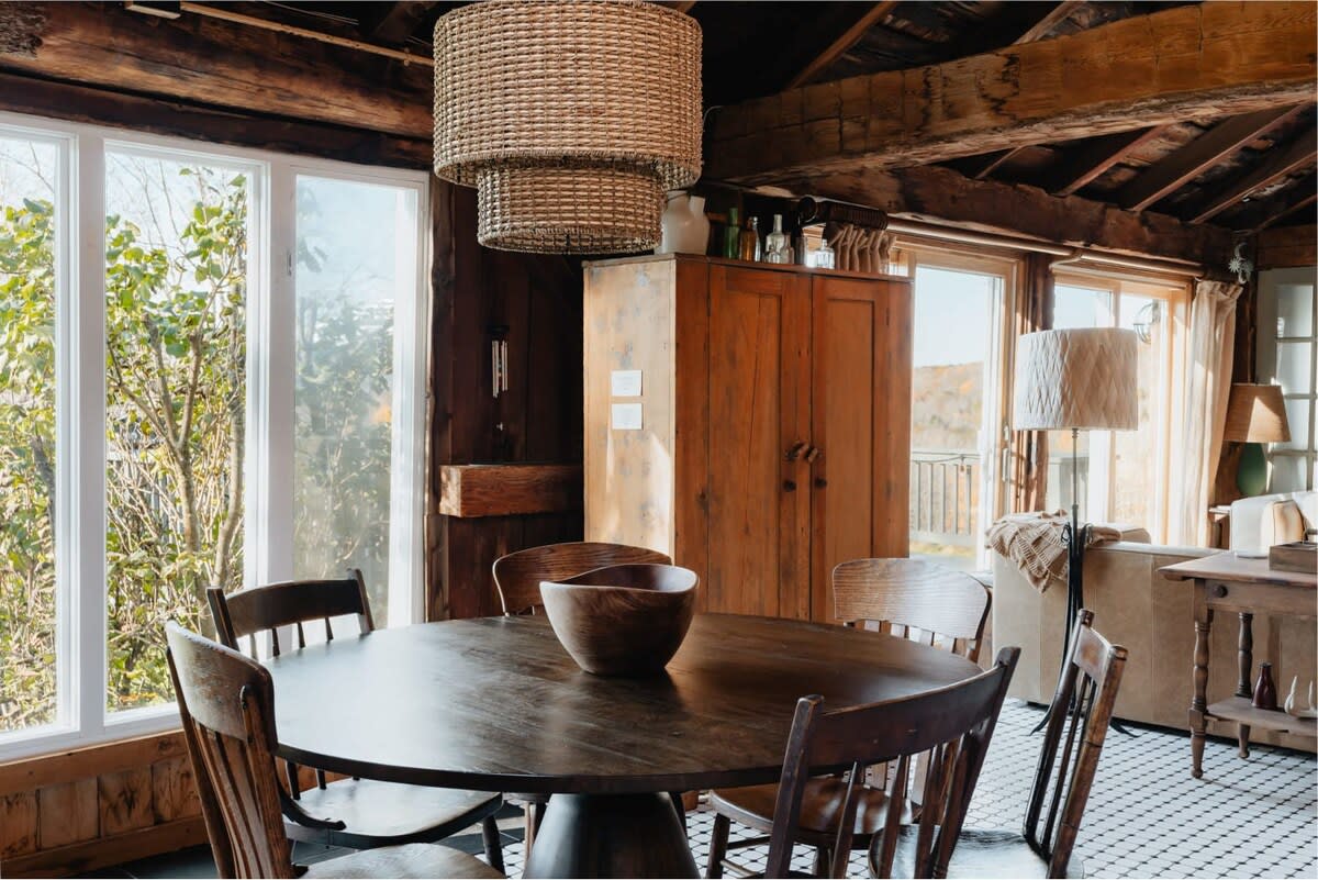 Rustic dining area featuring a round dark wood table with matching chairs, illuminated by a woven pendant light. Large windows offer views of the surrounding greenery.