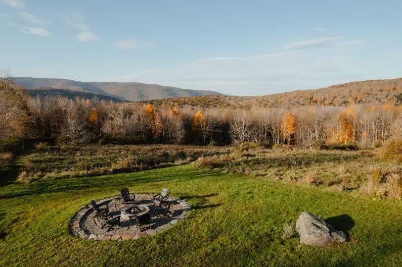 An aerial view of a stone-ringed fire pit area with four Adirondack chairs, set in a grassy field with rolling hills and autumn-colored forests in the background.