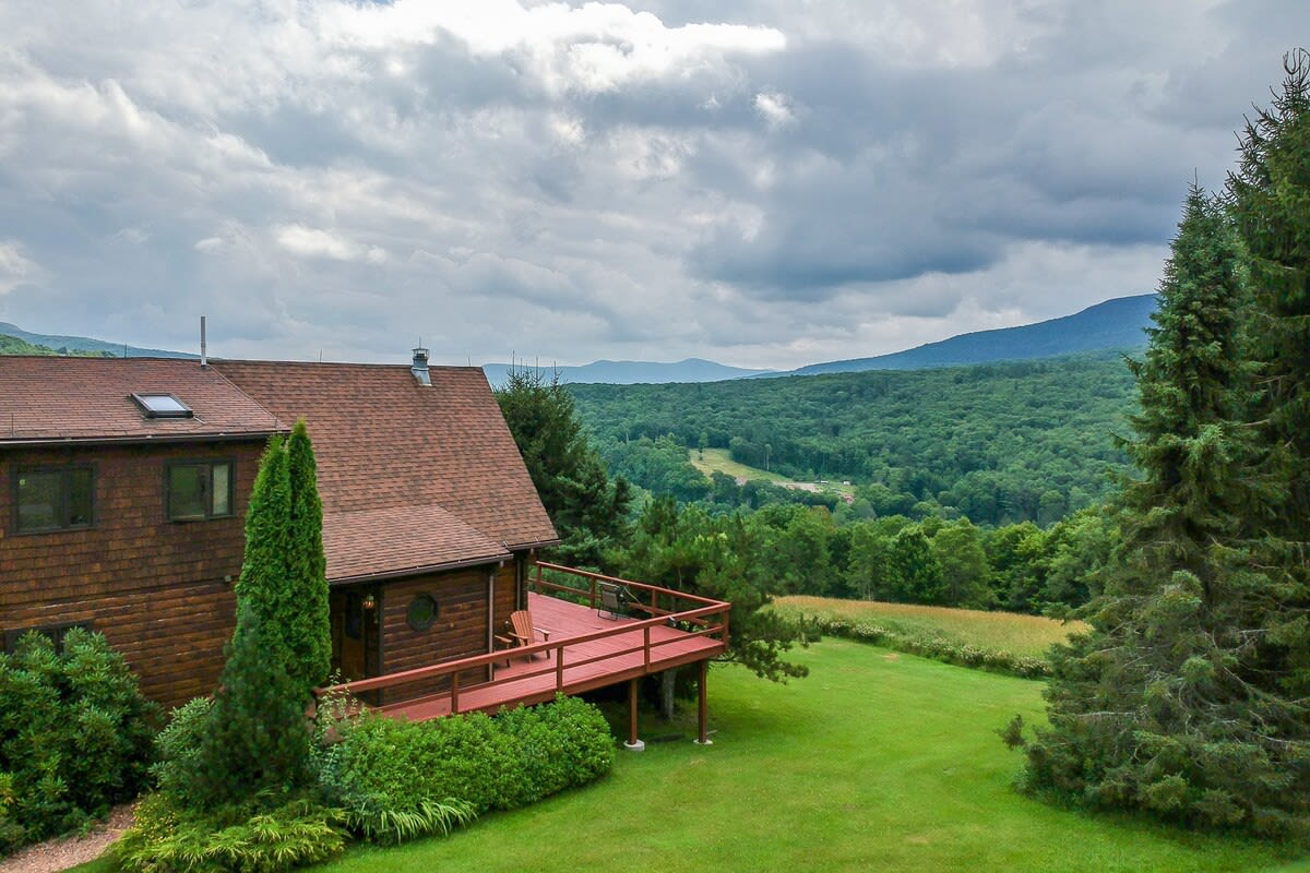 Elevated view of the side of a brown chalet-style house with a red roof and a large red-stained wooden deck, overlooking a vast green forest and distant mountains under a partly cloudy sky. Lush green lawn and tall evergreen trees frame the foreground.