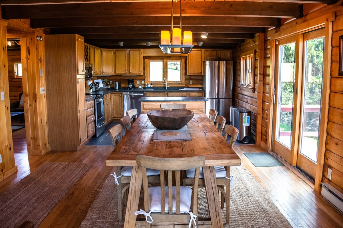 A well-appointed kitchen featuring light wood cabinets, dark countertops, and stainless steel appliances, with a large wooden dining table and chairs in the foreground.