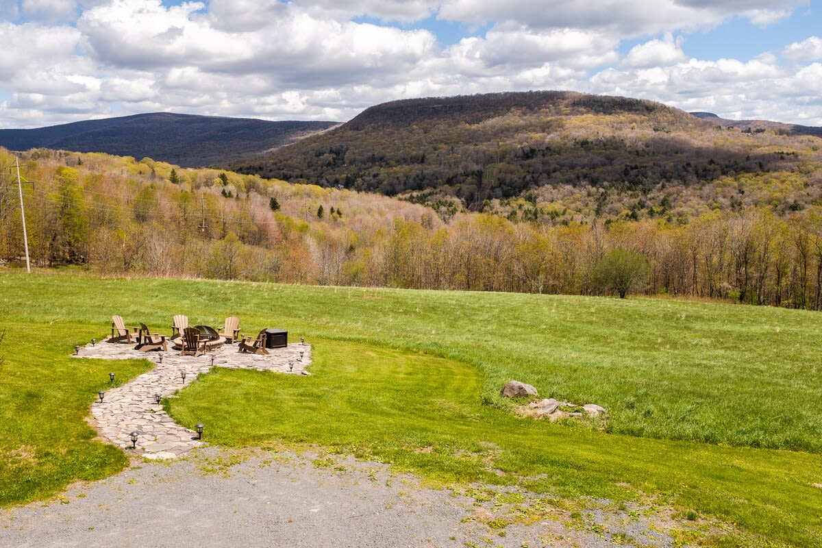 An aerial view captures a stone fire pit area with Adirondack chairs set in a sprawling green lawn, leading to distant rolling mountains under a partly cloudy sky. A stone path leads to the fire pit.