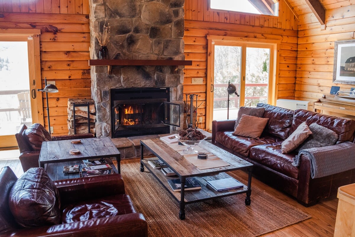 A rustic living room with log walls and a grand stone fireplace, furnished with comfortable leather sofas and industrial-style coffee tables. A warm fire glows in the hearth.