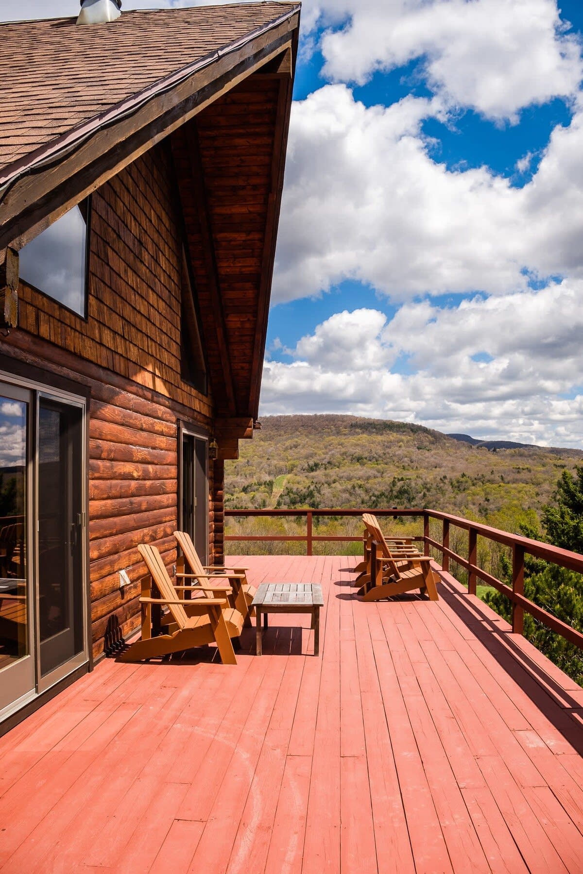 A wide red-painted wooden deck offers stunning mountain views under a blue sky with white clouds. Several wooden Adirondack chairs and a small table are arranged for relaxation.