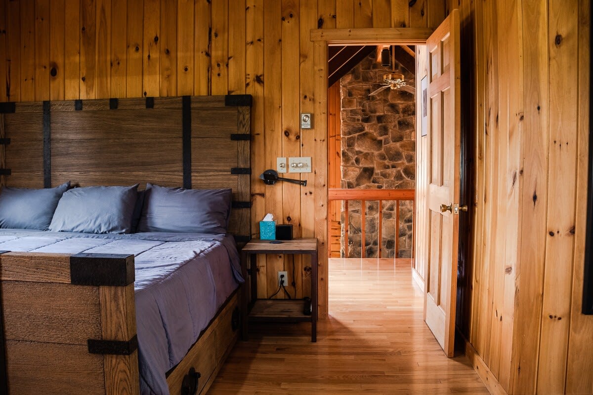 A rustic bedroom with warm wooden plank walls and a large wooden bed with a dark headboard and grey bedding. A doorway reveals a stone fireplace and a railing in an adjacent living area.