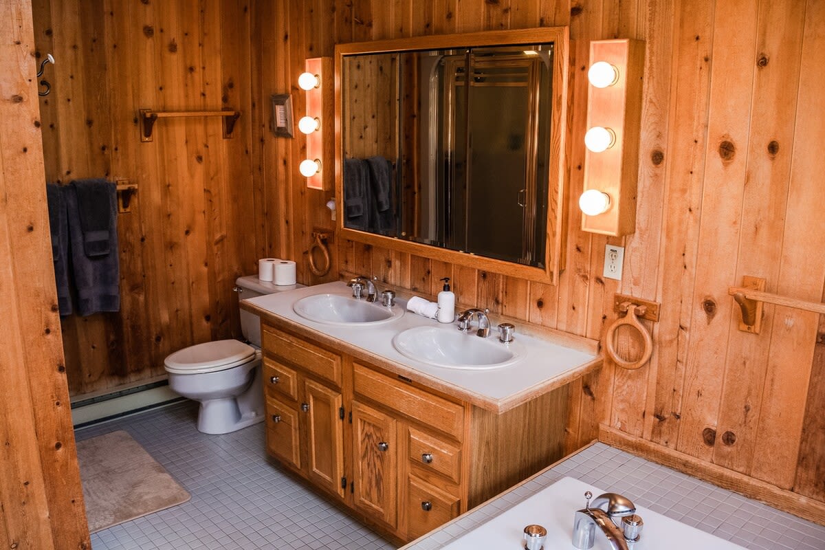 A rustic bathroom features warm wooden plank walls, a double vanity with white sinks, and a toilet. Bright vanity lights flank a large mirror, and a white bathtub is partially visible.