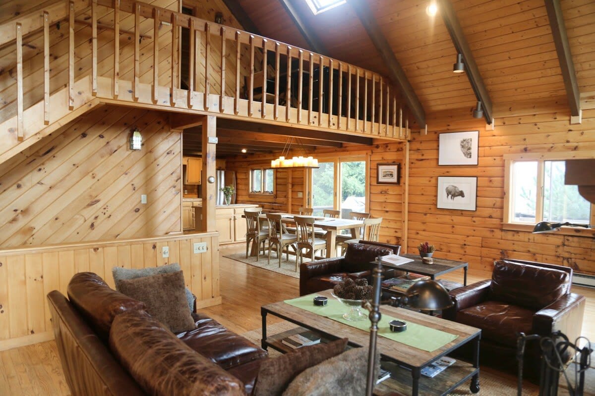 An open-concept living space with vaulted wood ceilings and log walls, featuring a living room area with leather seating, a dining area, and a glimpse into the kitchen.