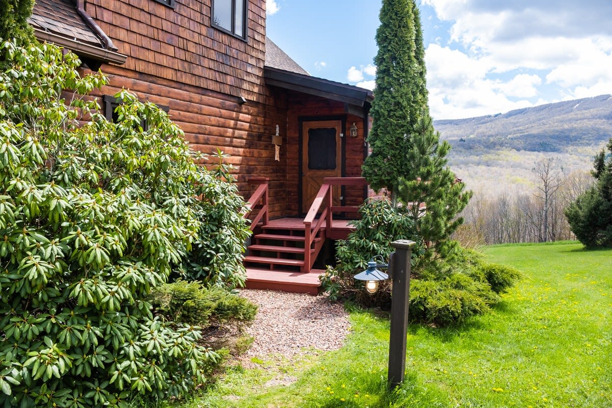 Close-up view of the main entrance to the brown chalet-style house, featuring a red-stained wooden porch with steps leading to a dark wood door. Lush green rhododendron bushes and a tall slender evergreen tree flank the entrance, with a rustic outdoor lamp post.