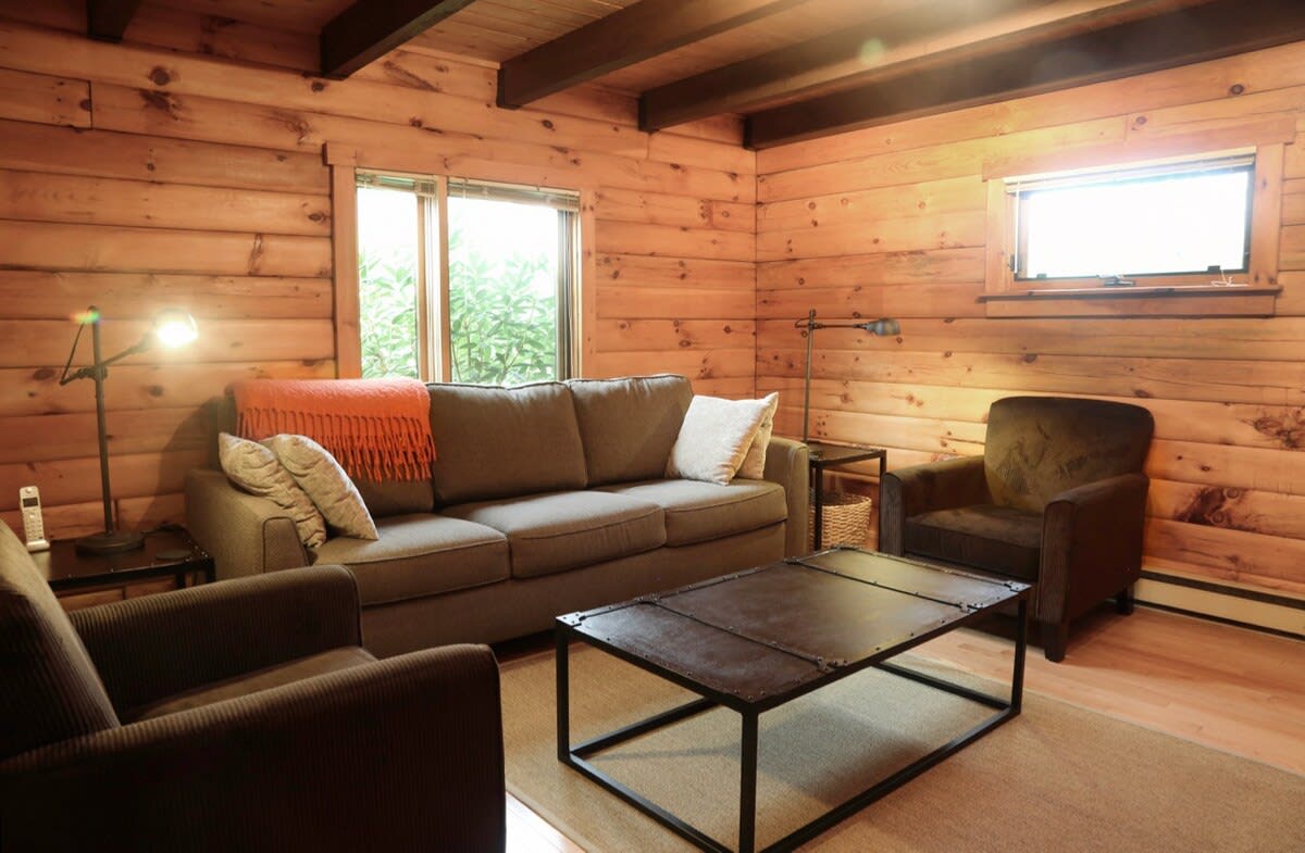 A cozy secondary living room with wood-paneled walls, furnished with a comfortable sofa, two armchairs, and an industrial-style coffee table. Windows offer views of surrounding greenery.