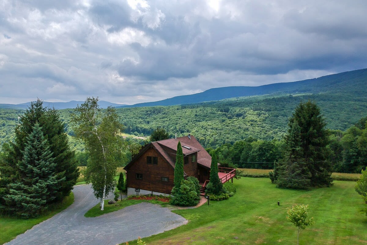 Aerial view of a brown chalet-style house with a red roof, nestled in a lush green landscape with dense forests and distant mountains under a cloudy sky. A gravel driveway curves towards the house, surrounded by mature evergreen trees and a birch tree.