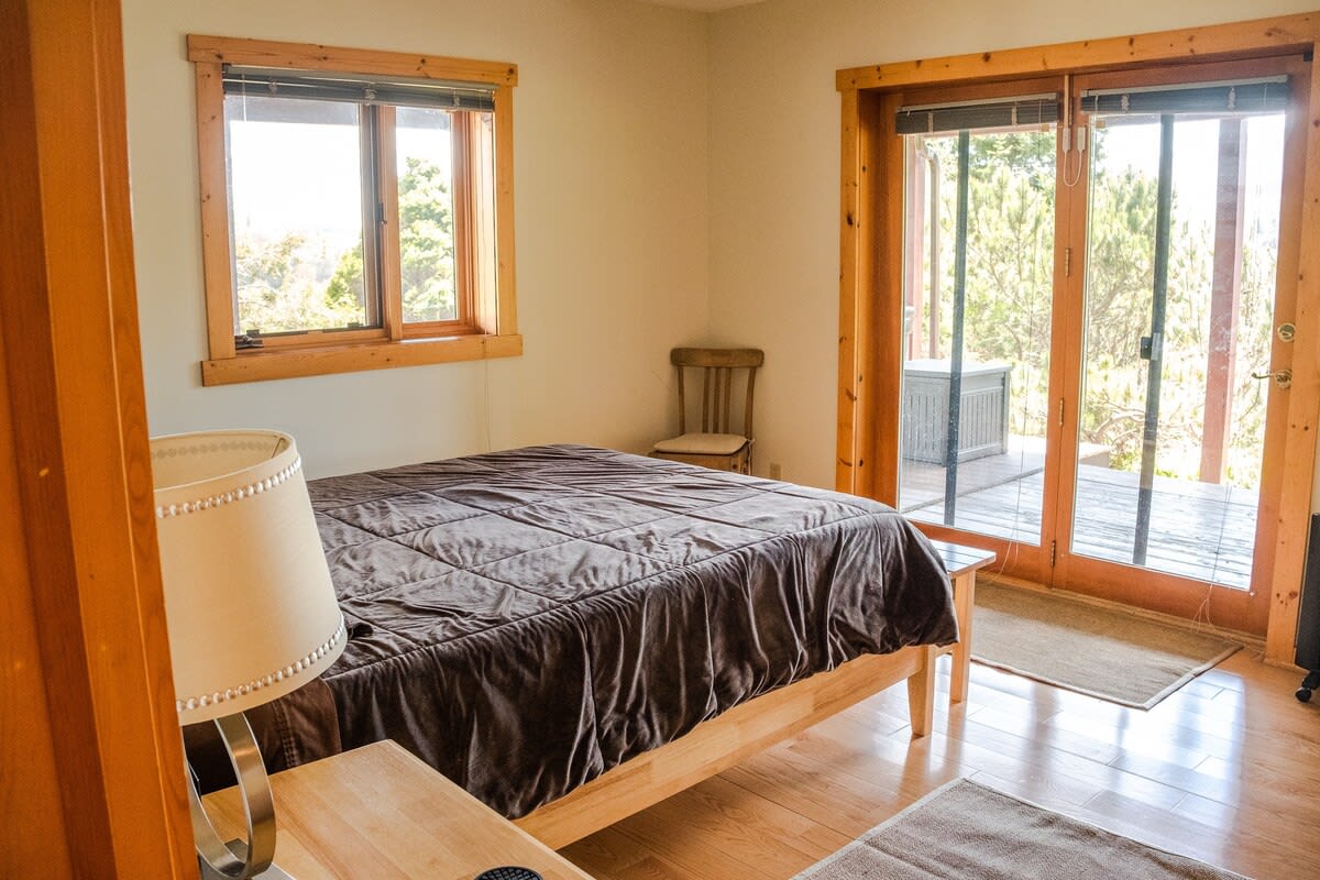 A serene bedroom features light walls and a light wooden bed frame with brown bedding. A large sliding glass door opens to an outdoor deck, providing natural light and a view of trees.