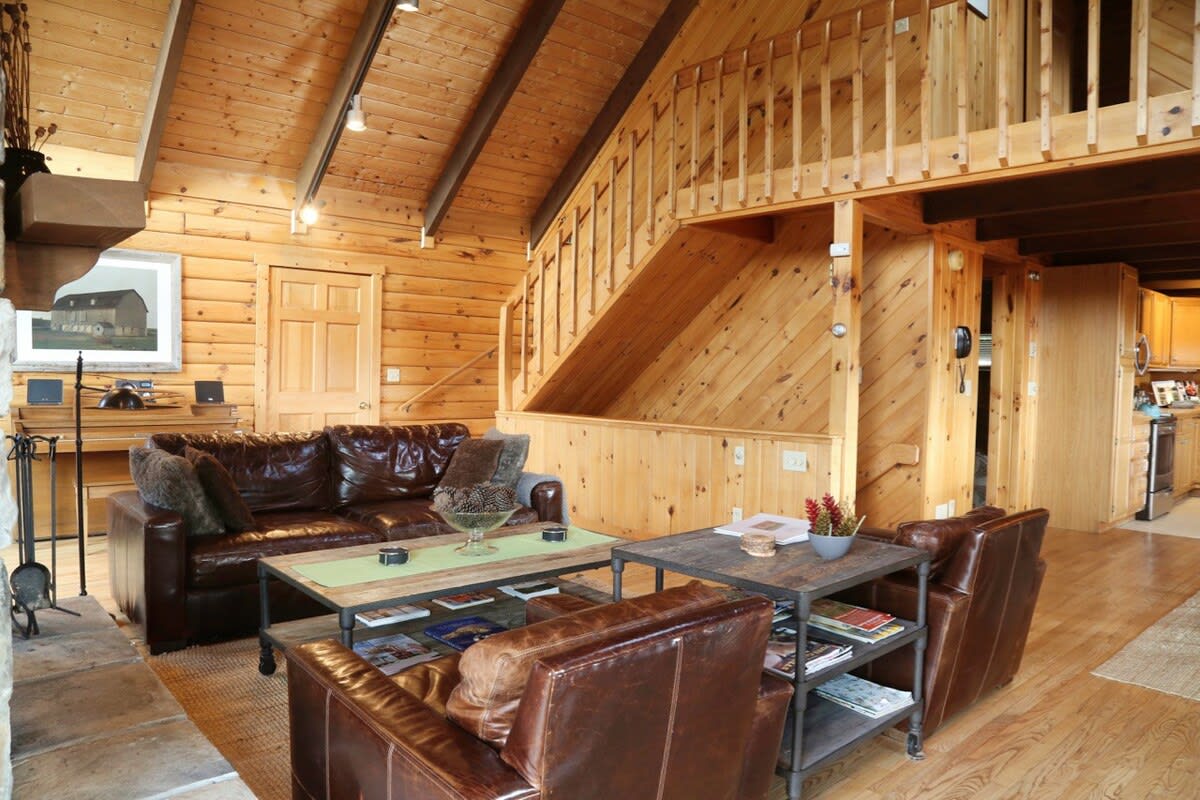 A spacious living room with warm wood-paneled walls and hardwood floors, featuring leather seating, a stone fireplace, and an open wooden staircase leading to an upper loft.