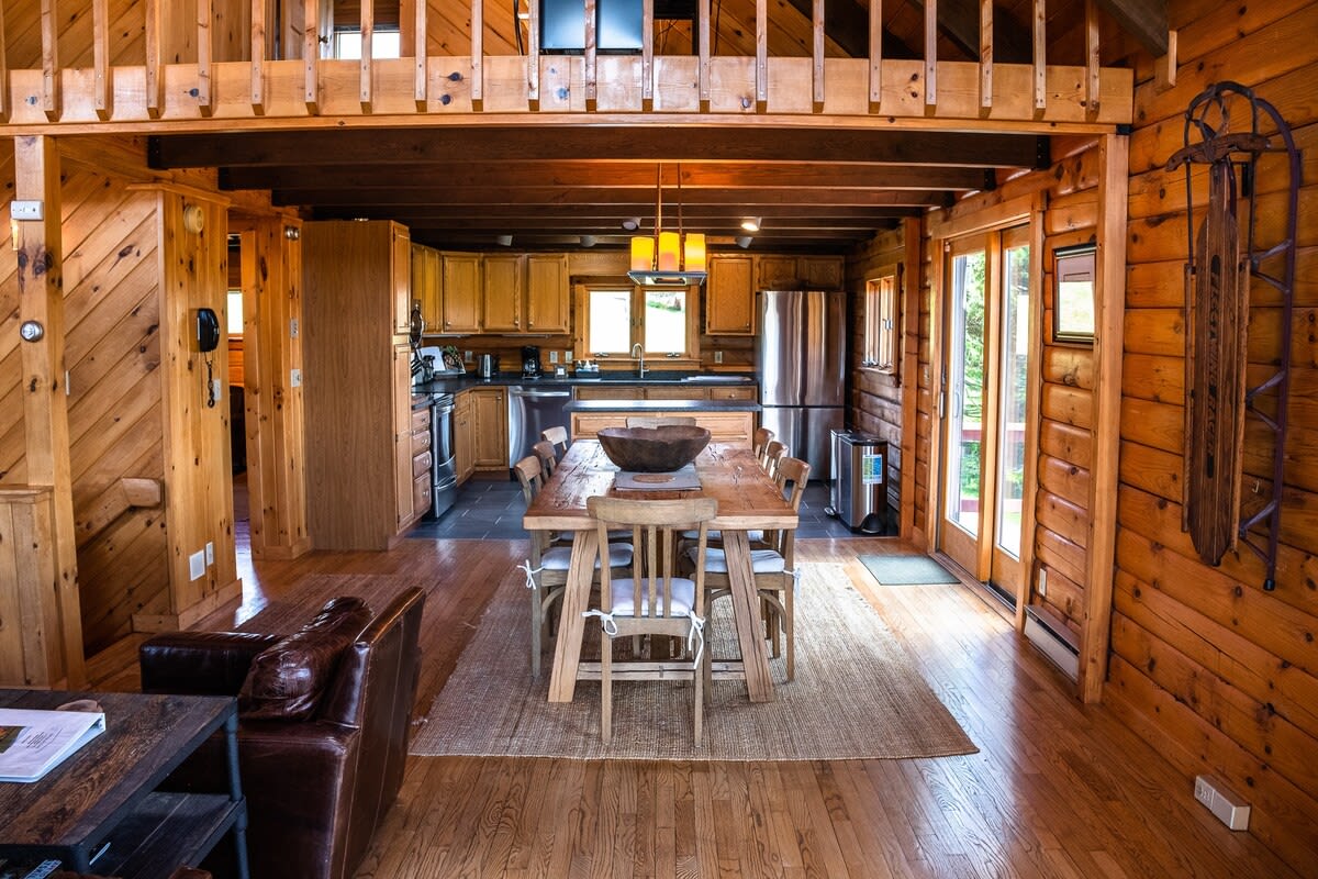 A rustic dining area featuring a large wooden table with chairs, situated within an open-concept space that includes a view of the adjacent kitchen with wooden cabinetry.