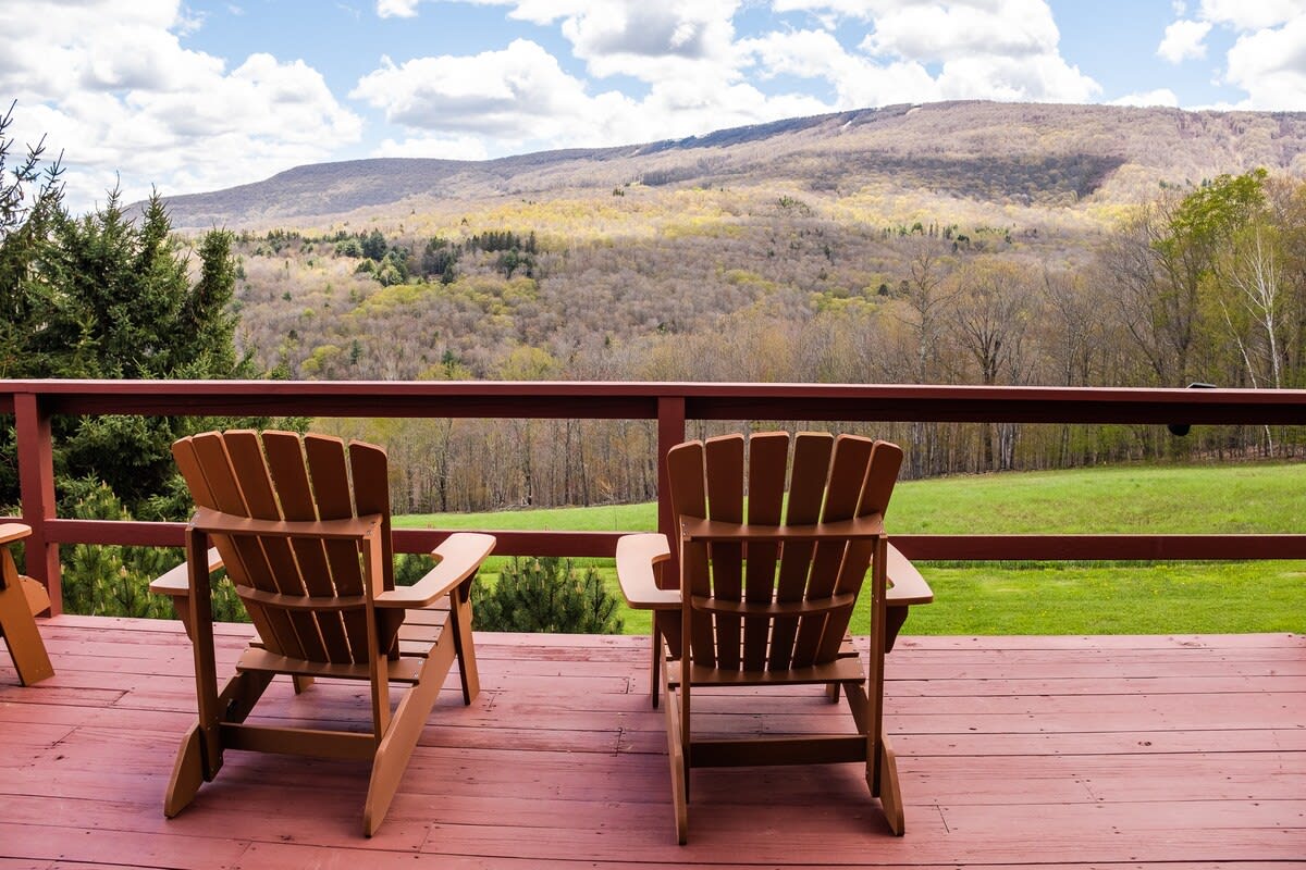 A spacious wooden deck furnished with two Adirondack chairs, offering a serene spot to enjoy expansive views of a forested mountain range under a bright, cloudy sky.
