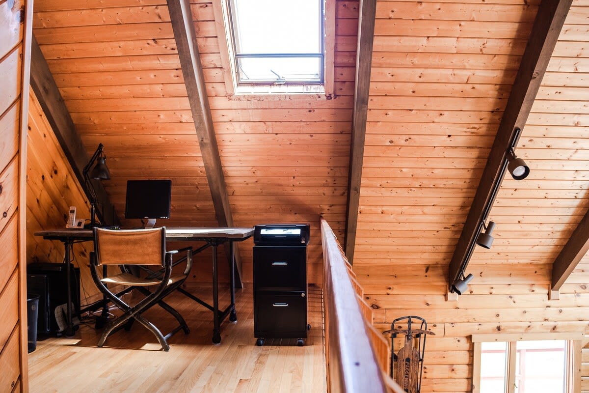 A cozy attic office space features a sloped wooden plank ceiling with exposed beams and a skylight, overlooking a desk with a computer and a black filing cabinet.