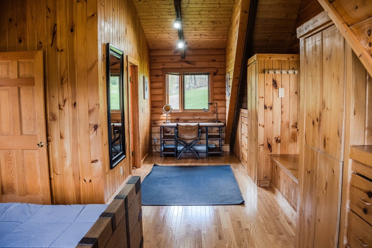 A cozy bedroom with light wood-paneled walls and hardwood floors, featuring a bed in the foreground and a dedicated workspace with a desk and chair by a window.