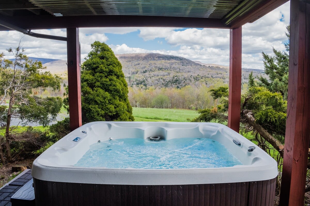 A bubbling hot tub is set on a covered outdoor deck, offering panoramic views of lush green mountains and a partly cloudy sky. Surrounding trees frame the scenic vista.
