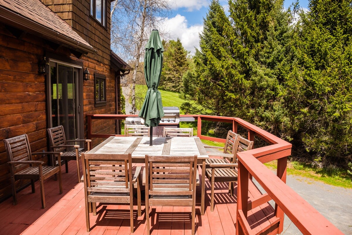 A spacious red-painted wooden deck features an outdoor dining set with an umbrella and a BBQ grill, surrounded by lush greenery and trees. The house exterior is visible on the left.