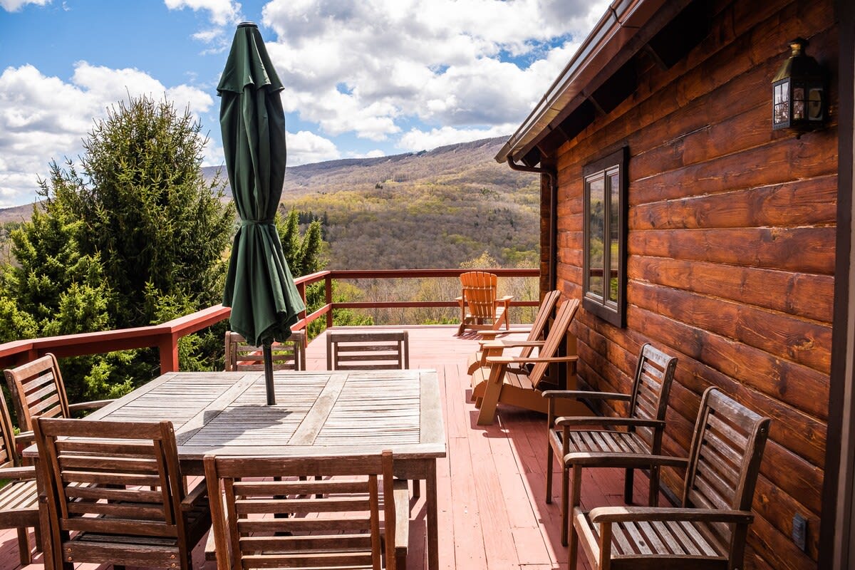 A spacious wooden deck attached to a log cabin, furnished with an outdoor dining set and lounge chairs, offering expansive views of a mountainous landscape.
