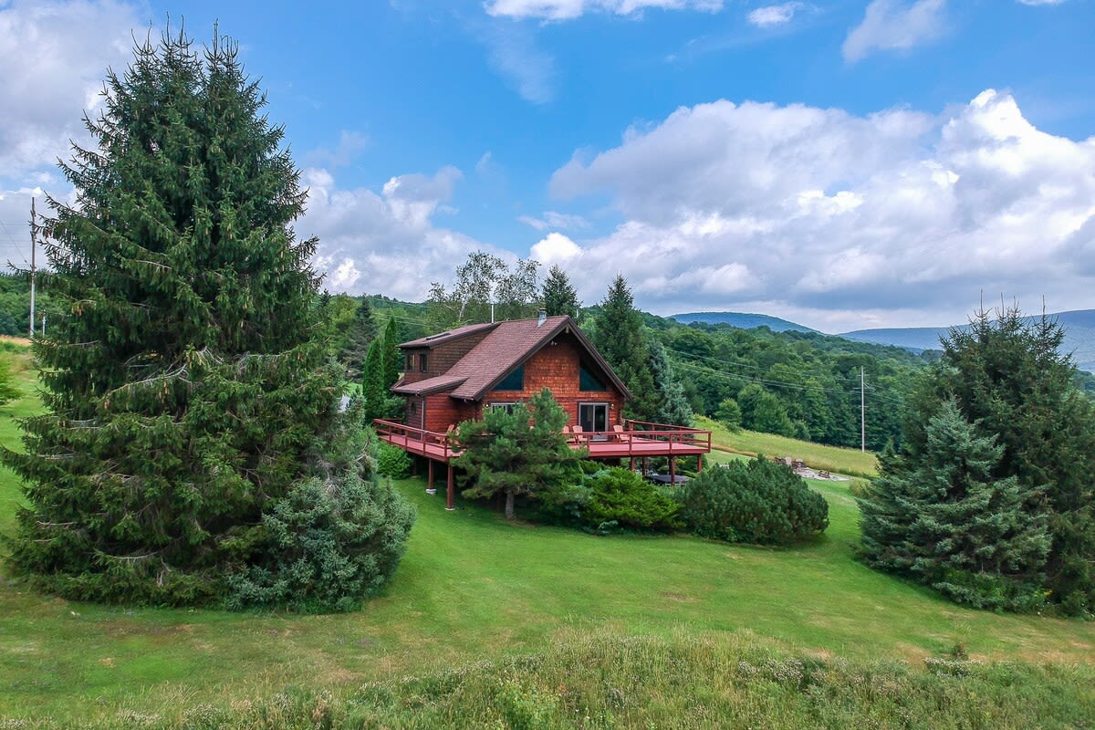 Wide exterior shot of a brown chalet-style house with a red roof and an expansive red-stained wooden deck, set on a grassy hill with tall evergreen trees. The background reveals rolling green mountains under a bright blue sky with white clouds.