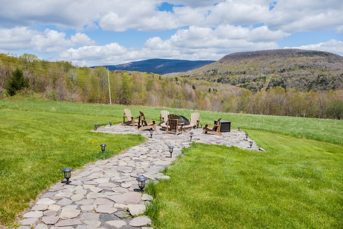 An inviting outdoor fire pit area with multiple Adirondack chairs arranged around a central fire pit, situated on a stone patio at the end of a flagstone path in a grassy field, with mountain views.