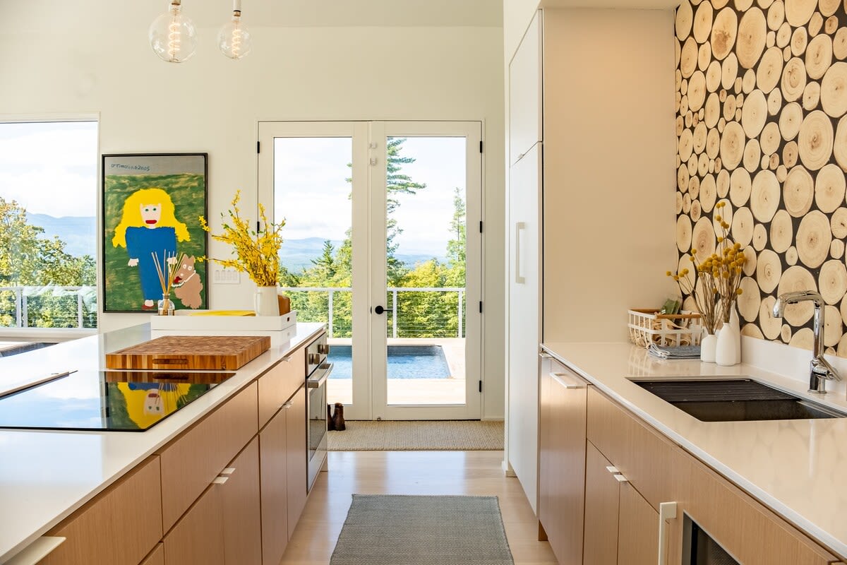 Modern kitchen with light wood cabinetry, white countertops, and a unique accent wall made of stacked wood slices, featuring a built-in stovetop and a sink, with French doors leading to an outdoor area with a pool and mountain views.