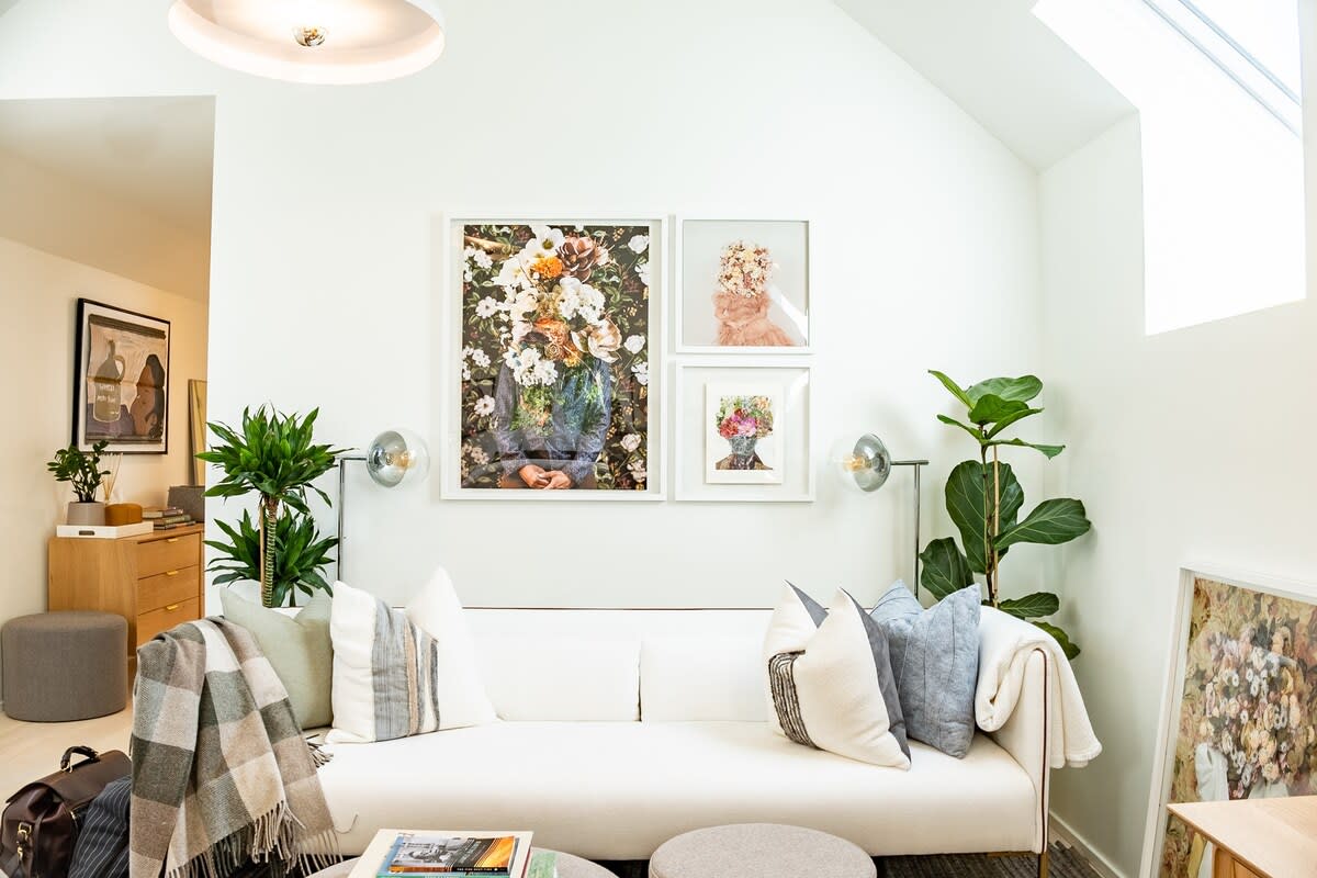 A bright living room with a white sofa adorned with various pillows and a plaid throw, featuring a gallery wall of floral-themed artworks, and large windows allowing natural light. Potted plants add a touch of greenery.